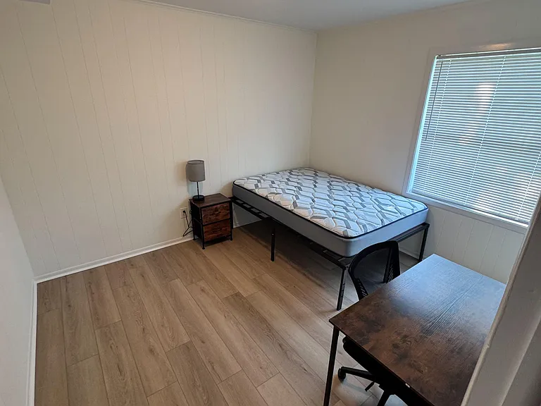 Furnished bedroom with wood-style floors, a bed frame and nightstand, plus a dedicated desk and window with blinds.