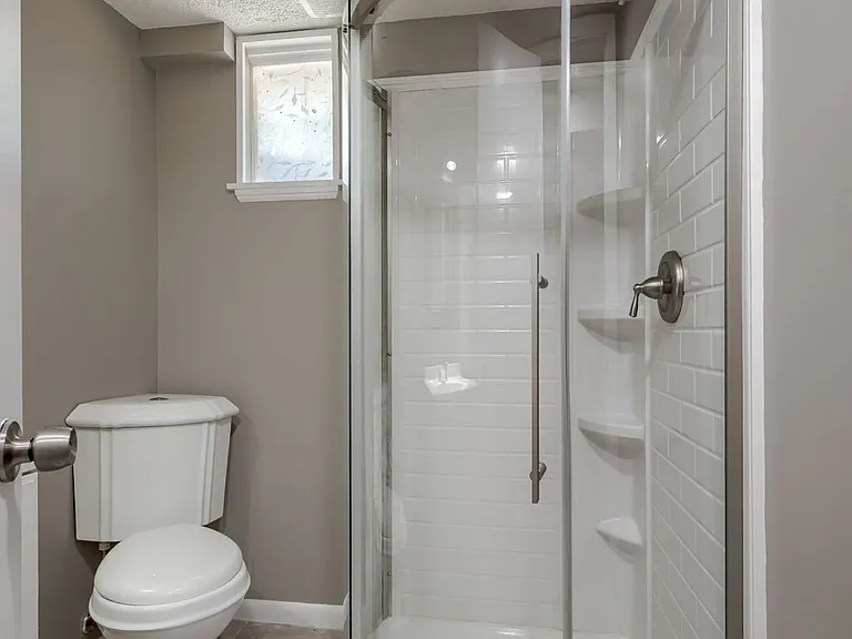 Bathroom3: Primary downstairs bathroom featuring corner toilet, shower with overhead showerhead, and small tinted window into the backyard