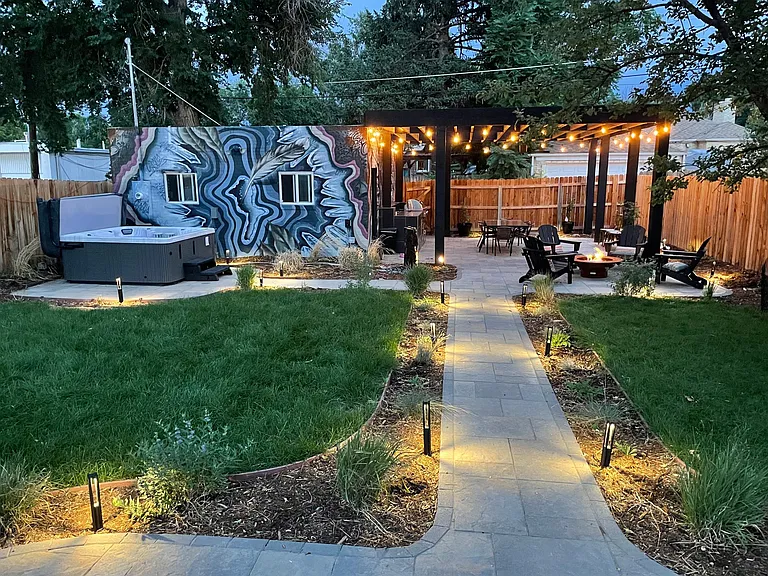Backyard at night with beautiful flower gardens, outdoor fire pit, jacuzzi, kitchen and covered dining area.