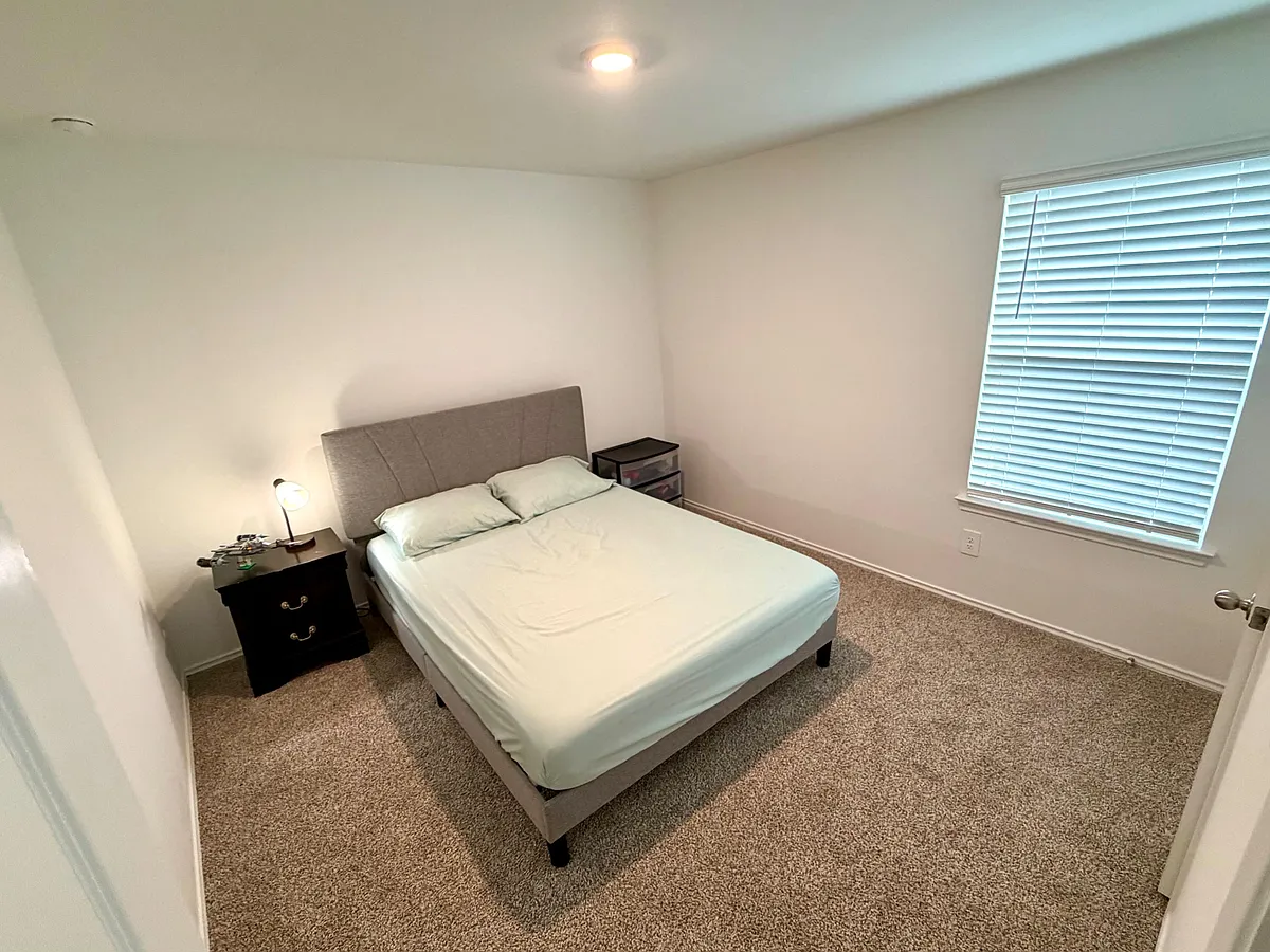 Furnished bedroom with a full bed, nightstand, and dresser, plus a large window with blinds and carpeted flooring. - Eastern Pine Trl