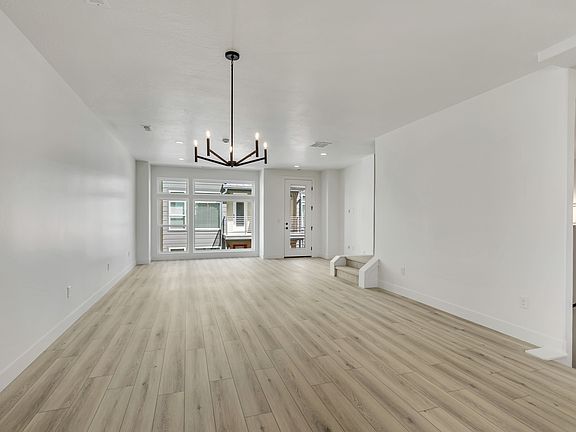 A modern empty living room with light wood flooring, white walls, a black chandelier, large windows,