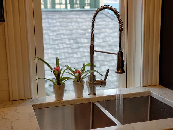 Nice light from a long window over a deep stainless steel sink with modern fawcet with two spraying options. White solid quartz countertop with random black design.