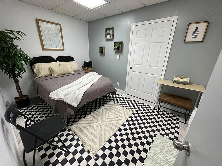 Furnished private bedroom featuring a full bed, accent rug, wall shelving, and a small desk/bench area for daily use.