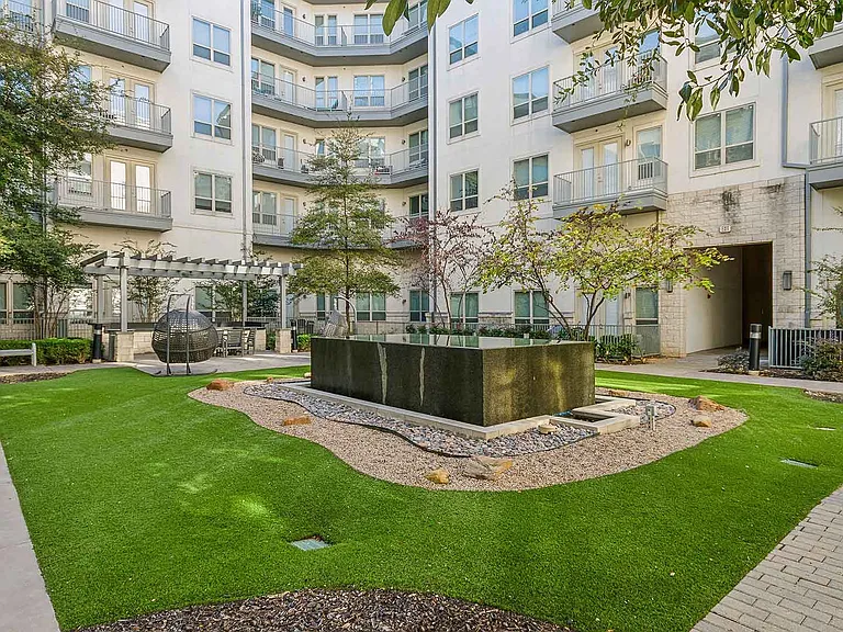 A tranquil courtyard in a residential complex, featuring a modern water fountain surrounded by decorative pebbles and vibrant green artificial turf. The space includes a pergola with outdoor seating a