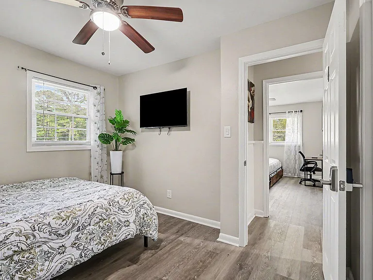 Furnished bedroom with a ceiling fan, large window with blinds, and wall-mounted TV, plus easy access to the hall.