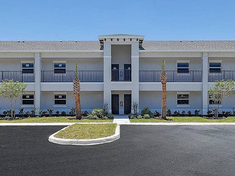 Modern two-story apartment building with elegant balconies, surrounded by lush landscaping and clear blue sky.