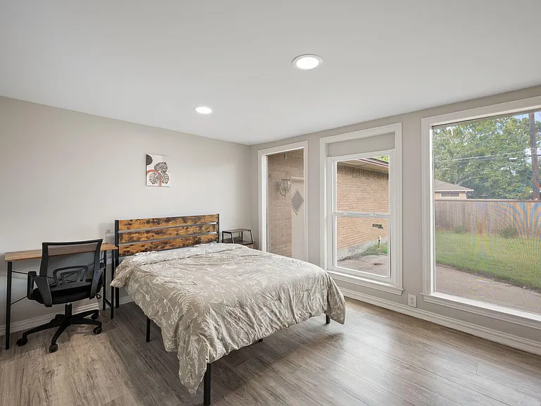 Furnished bedroom with a work desk, ceiling lighting, and large windows overlooking the yard.