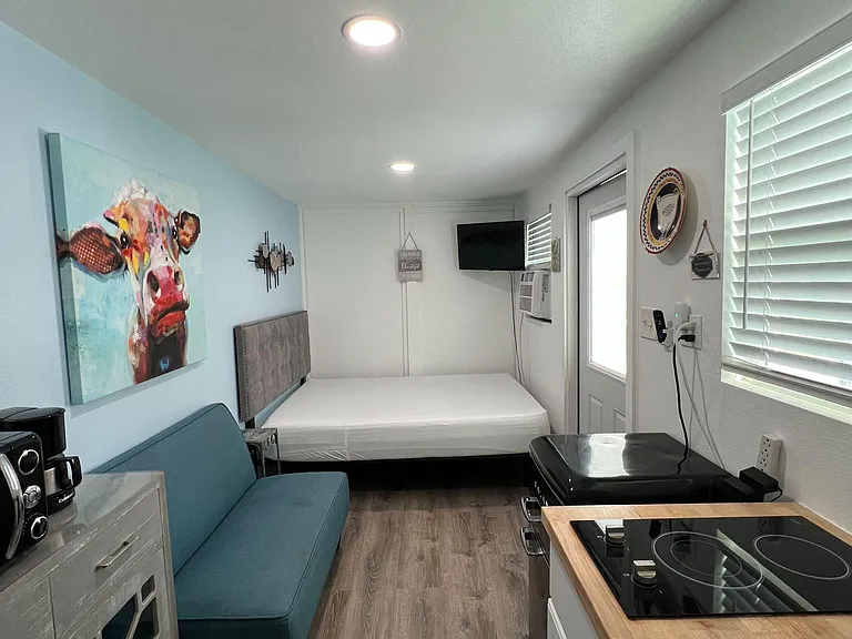 Efficient studio-style room with kitchenette, seating area, and sleeping space, plus wall-mounted TV and window blinds.