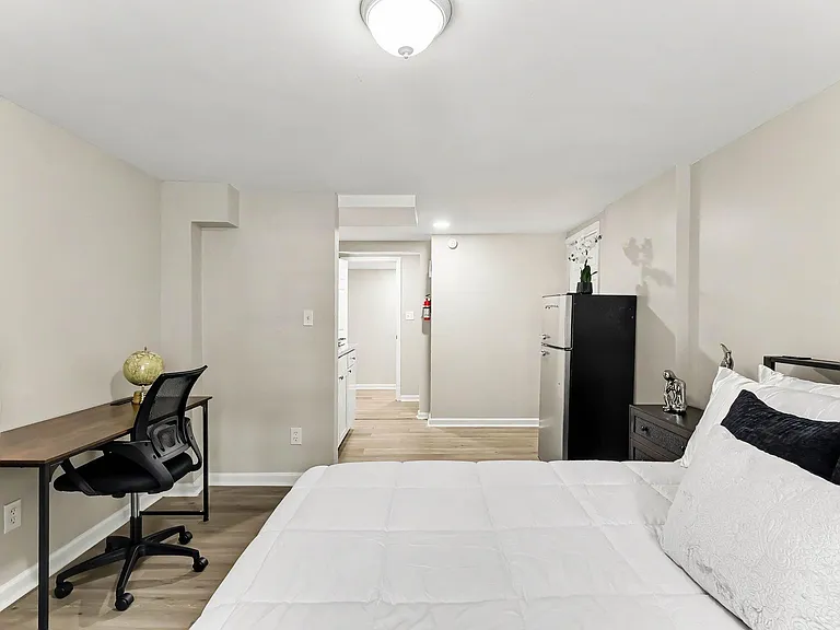 Spacious bedroom setup with a dedicated desk area, mini fridge, and easy access to the adjoining hallway and bath.