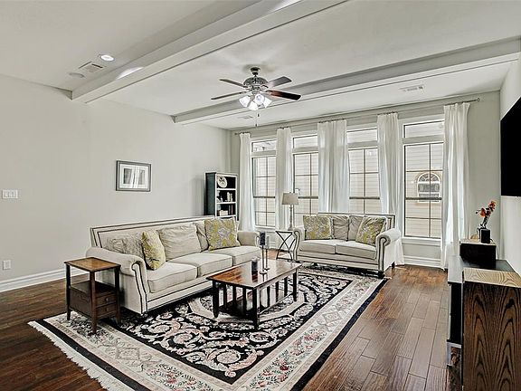 This bright and spacious living room has a high ceiling with decorative beams and ceiling fan, gleaming wood floors and an impressive wall of windows to let in the natural light.