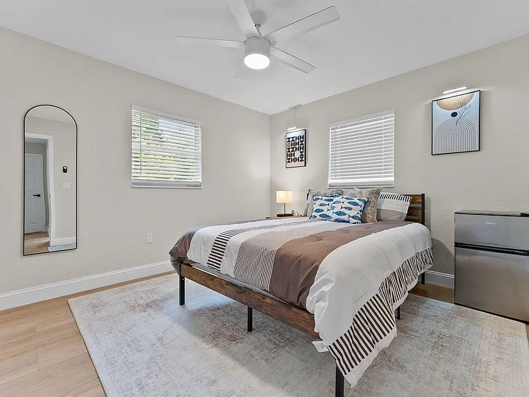 Well-sized bedroom with a queen bed, ceiling fan, two windows with blinds, and space for added storage or a mini fridge.