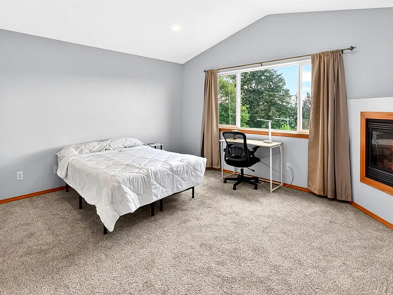 Spacious bedroom with vaulted ceiling, large window, dedicated desk area, and a built-in fireplace for added comfort.