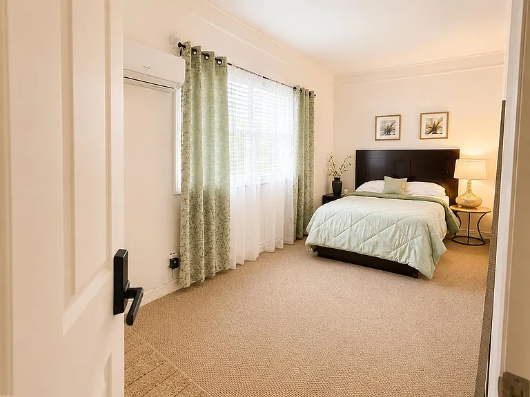 Spacious bedroom with a large window, blackout-style drapes, and a comfortable bed setup with nightstand lighting.
