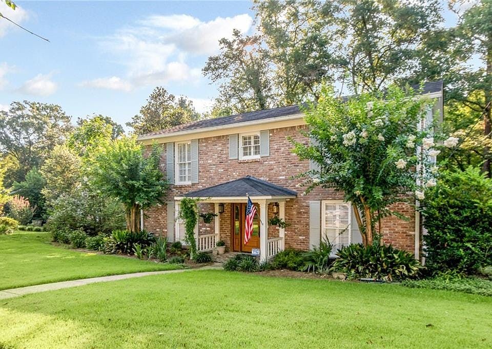 Brick facade residence featuring a covered front porch with white railing, mature landscaping, and an expansive front lawn