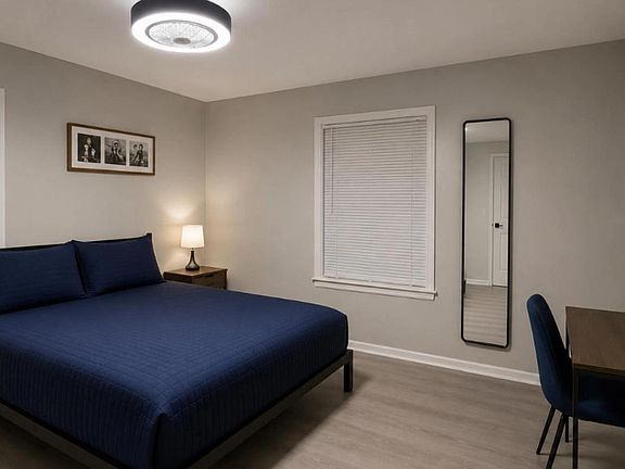 Spacious furnished bedroom with a full bed, nightstand lighting, dedicated desk area, and two windows with blinds.
