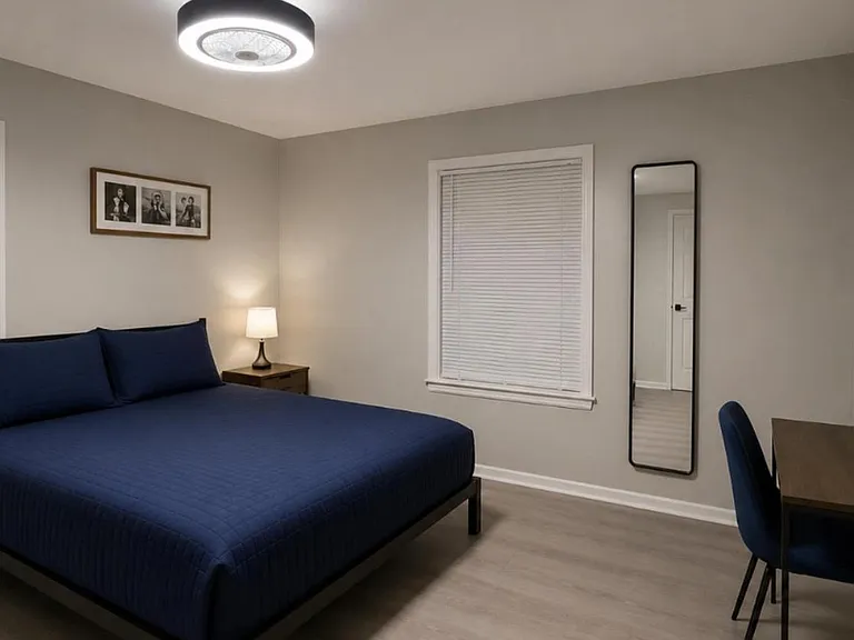 Spacious furnished bedroom with a full bed, nightstand lighting, dedicated desk area, and two windows with blinds.