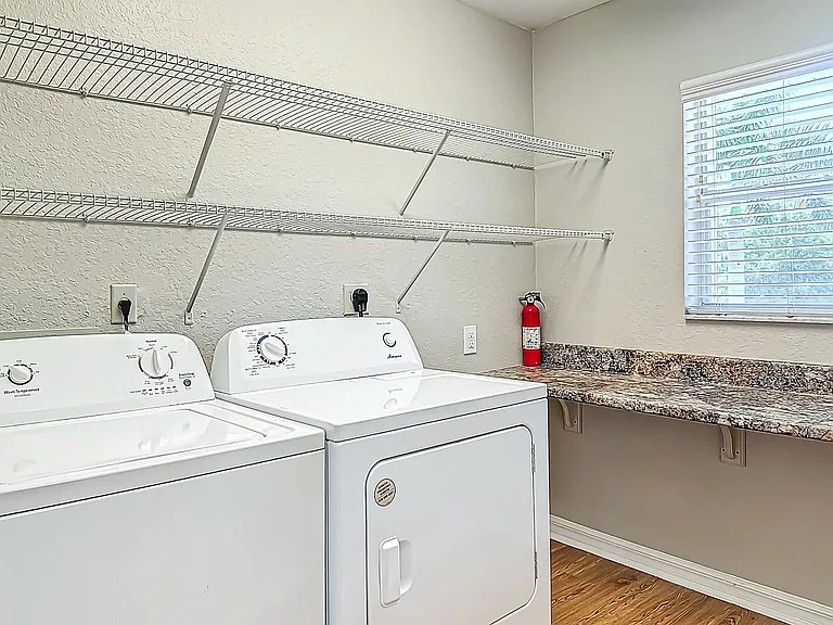 This in-unit laundry room offers ample space for all your laundry needs, featuring a washer, dryer, built-in shelving, and a large countertop beneath a bright window.