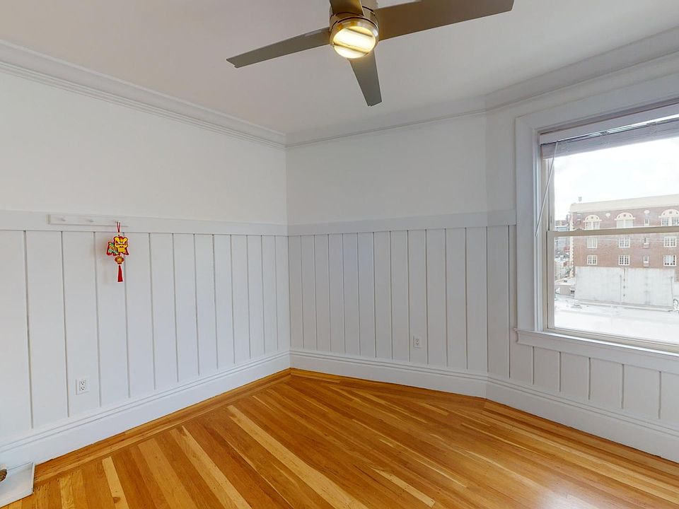 Bright corner room with wainscoting, hardwood floor, ceiling fan, and a window overlooking a brick building, decorated with a lucky charm.