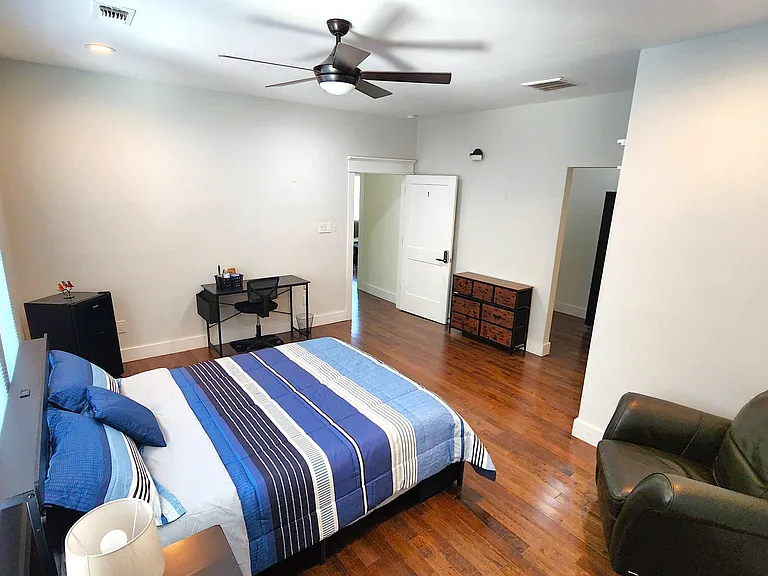 Spacious furnished bedroom with hardwood floors, ceiling fan, large window blinds, and room for a desk and seating.