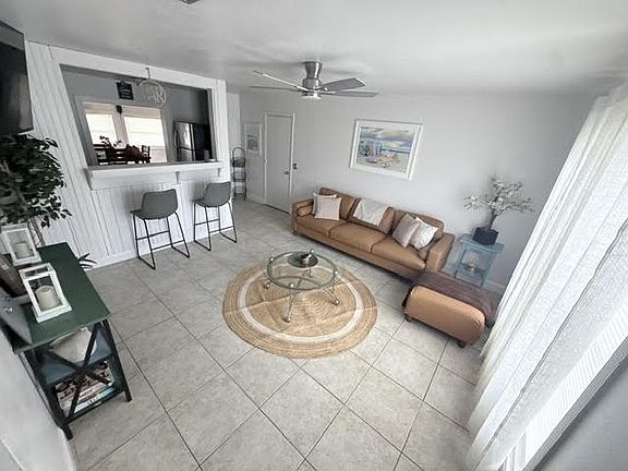Open living area with tiled floors, ceiling fan, and a seating layout anchored by a glass coffee table and large windows.