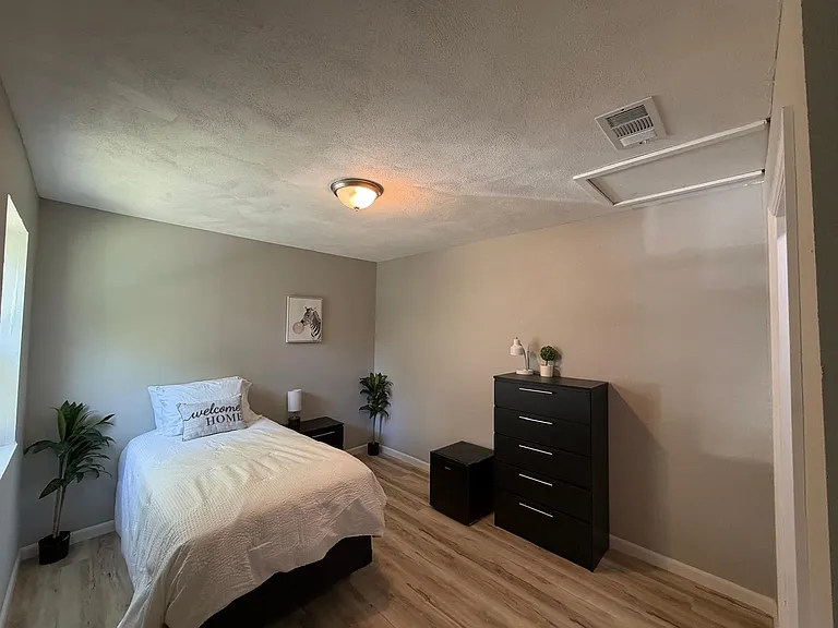 Furnished bedroom with a comfortable bed, dresser, mini fridge, and hardwood-style floors for easy upkeep.