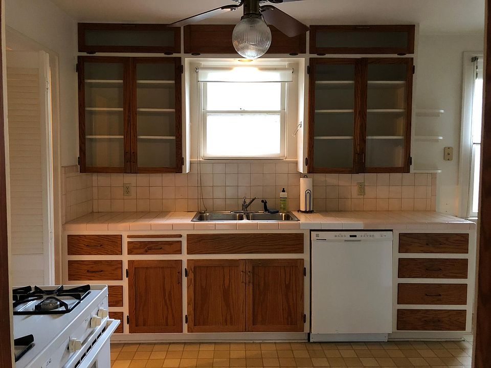 View of kitchen cabinets, sink, window above sink, dishwasher, natural gas stove and tiled backsplash.