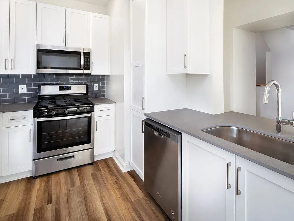 Kitchen with white cabinetry, grey quartz countertops, grey tile backsplash, stainless steel appliances, and hard surface flooring - Avalon at the Hingham Shipyard