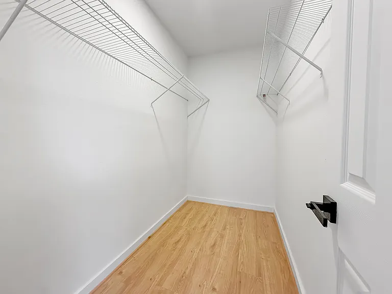 This photo shows a spacious walk-in closet with light wood flooring and white walls, featuring wire shelving on both sides for ample storage.
