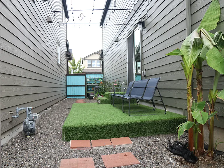 Cozy side_yard retreat with river rock pea gravel walkway, artificial_grass seating area, string lights, tropical greenery, and a colorful gate accented with potted plants.