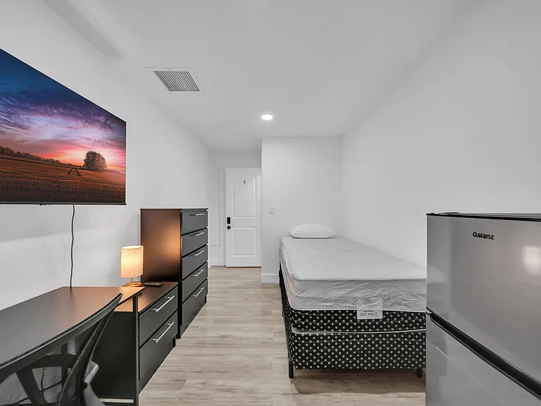 Furnished private bedroom with twin bed, work desk, dresser storage, wall-mounted TV, and an in-room mini fridge.