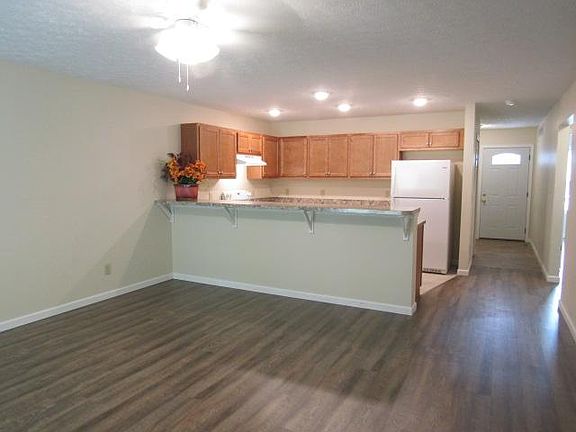 Open floor plan shows entry door, kitchen, living room with cream colored walls & white time, brown/gray vinyl tile transitions to faux wood brown laminate floor, breakfast bar with 4 supports. Photo shows ceiling fan with 4 lights, and 4 spotlights in k