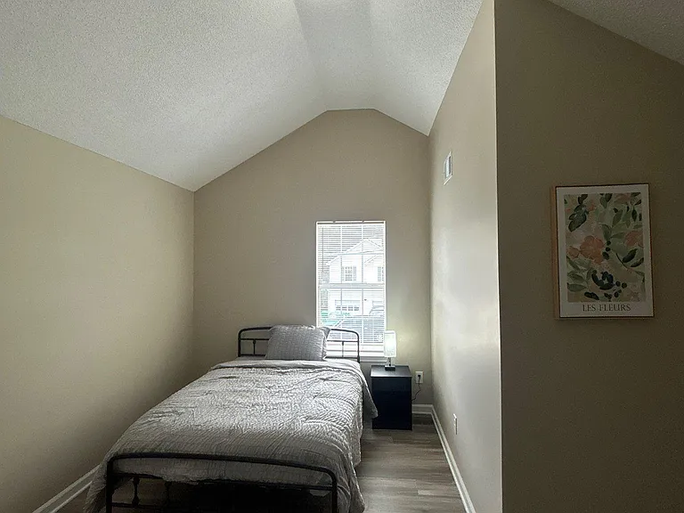 Private bedroom with vaulted ceiling, window with blinds, and durable flooring; furnished with bed and nightstand.