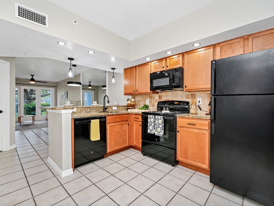 This renovated kitchen features modern black appliances, granite countertops, wood cabinetry, and pendant lighting. The open layout flows into a bright living space with tile flooring and French doors leading to the patio.