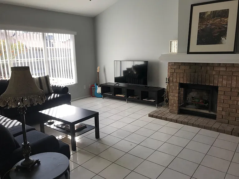 Spacious living room with tile flooring, large window with vertical blinds, and a brick fireplace for added character.