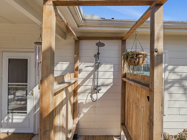 Outdoor shower enclosure with natural wood framing and slatted privacy walls, featuring a multi-function shower system and white horizontal siding backdrop