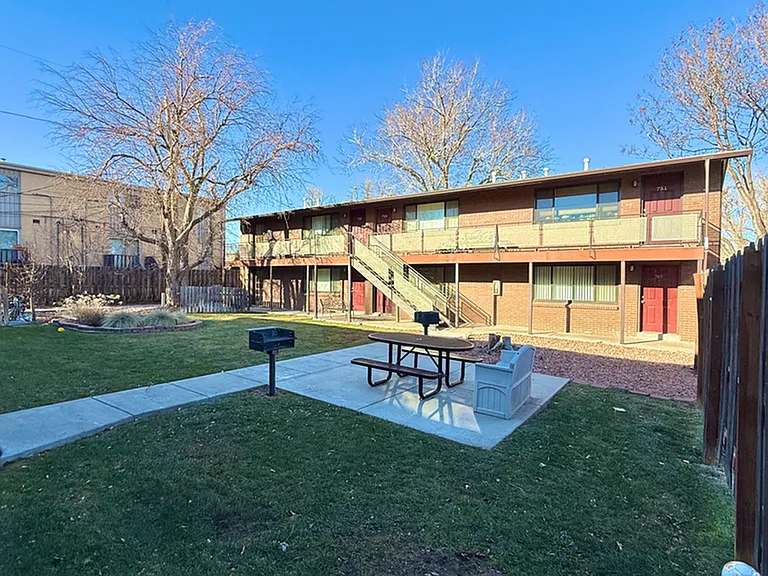 Cozy apartment complex with charming red doors and an inviting courtyard featuring a picnic area and barbecue spot, surrounded by lush greenery.