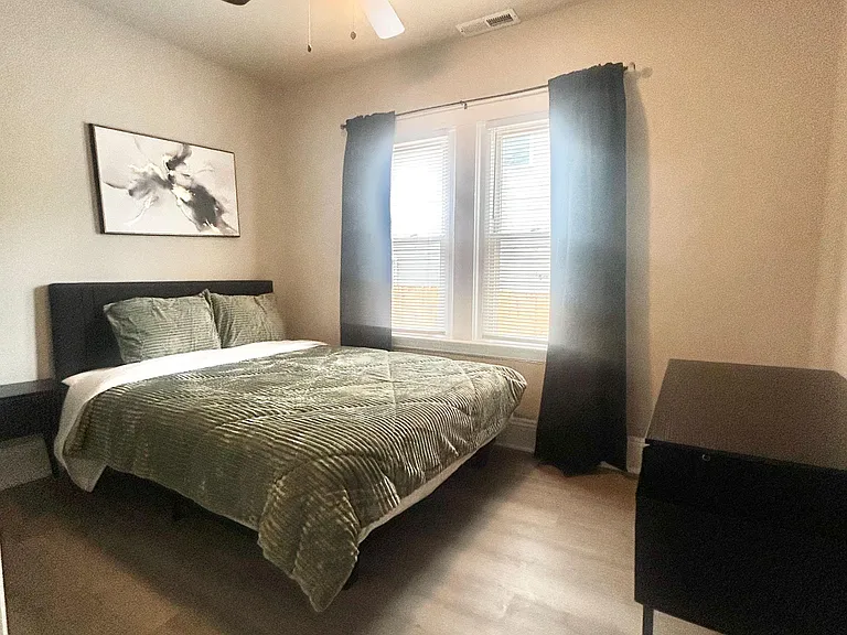 Furnished bedroom with a ceiling fan, large window with curtains, and space for a desk or dresser.