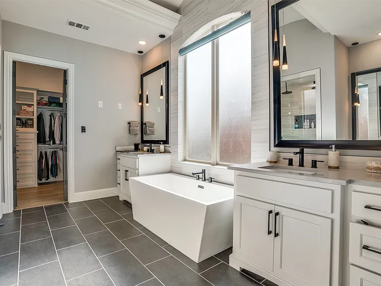 Spacious bathroom featuring a freestanding soaking tub, dual vanity with quartz countertops, custom cabinetry, and large format dark tile flooring