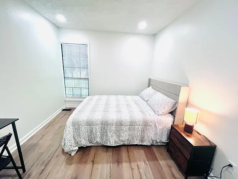 Private bedroom featuring a furnished bed, nightstand with lamp, and a window with blinds for light and privacy.