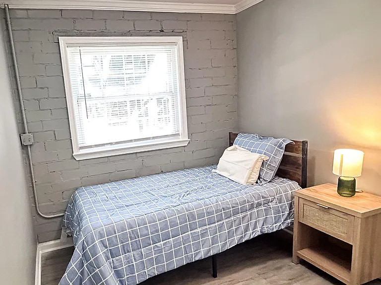Private bedroom with a single bed, nightstand, and overhead light, plus a window with blinds and durable flooring.