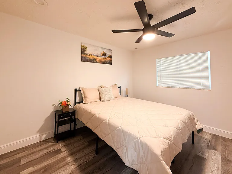 Furnished bedroom with a ceiling fan, window with blinds, and bedside tablecomfortable layout with wood-look flooring.