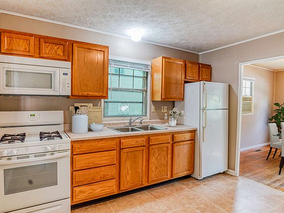 A simple kitchen featuring wooden cabinets, a gas stove, microwave, and refrigerator, with a window letting in natural light.