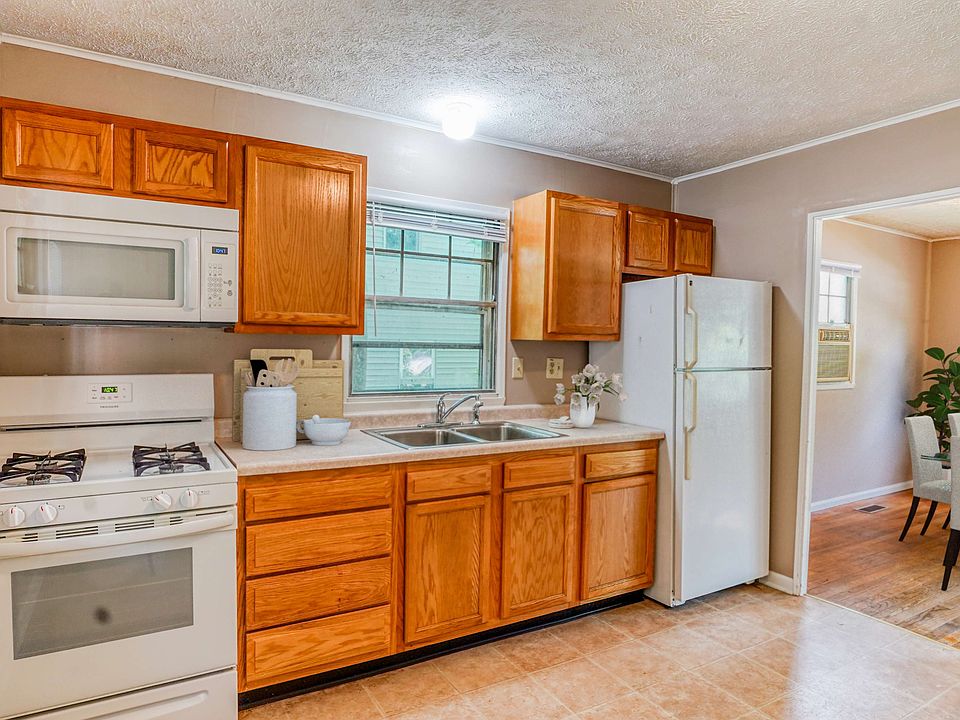 A simple kitchen featuring wooden cabinets, a gas stove, microwave, and refrigerator, with a window letting in natural light.