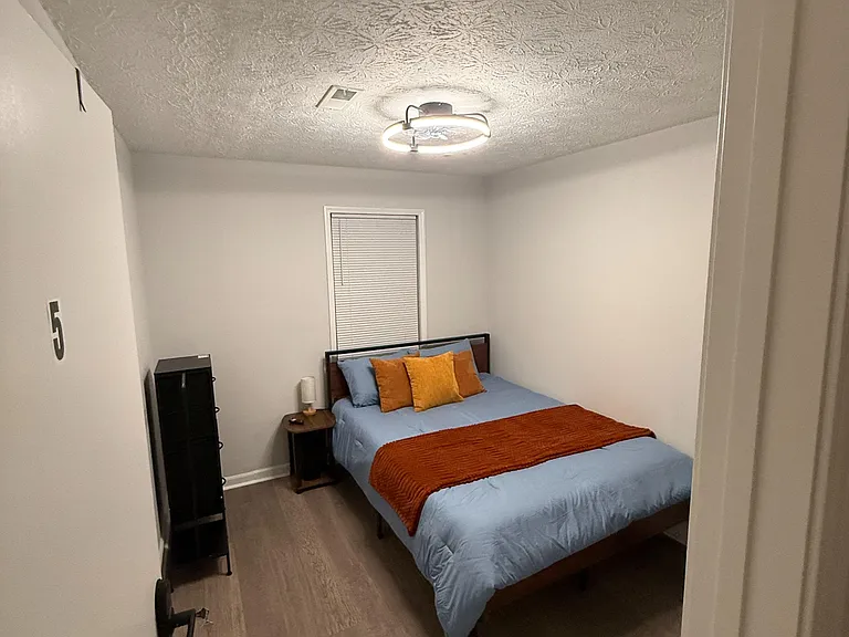 Private bedroom with a full bed, nightstand, and dresser, plus a window with blinds and overhead lighting.