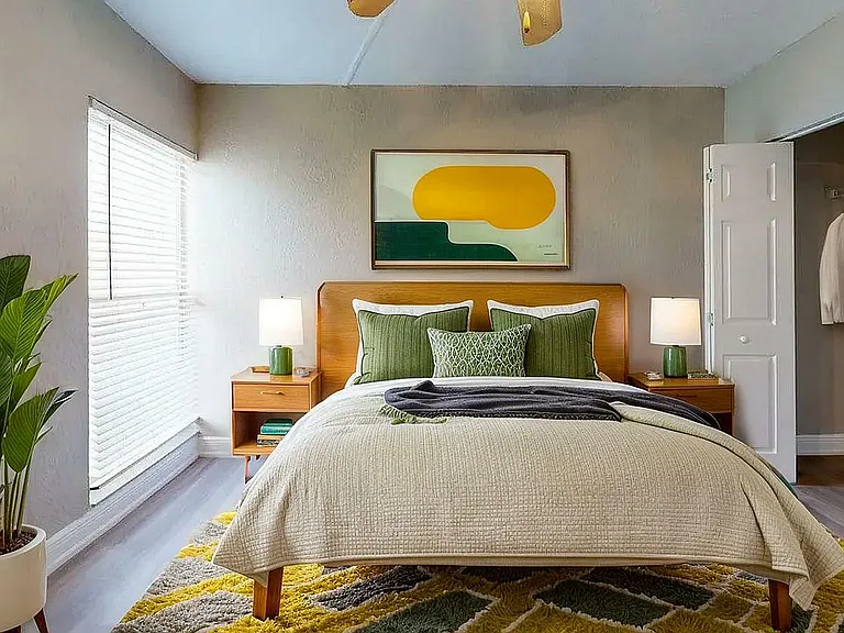 Bright and inviting bedroom with a modern aesthetic, featuring a plush bed with green accents, stylish artwork, vibrant rug, and ample natural light.