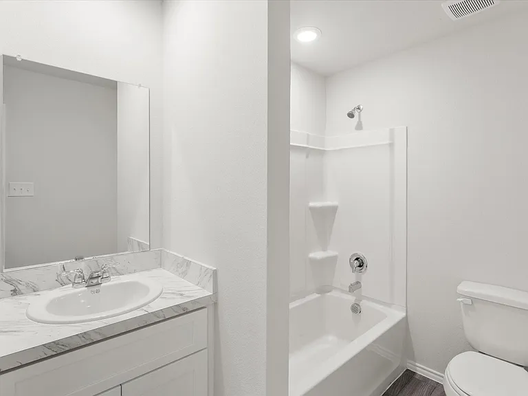 Bathroom featuring a white vanity with an integrated sink, a marble-patterned countertop, a mirrored wall, a white bathtub with a shower, and a white toilet