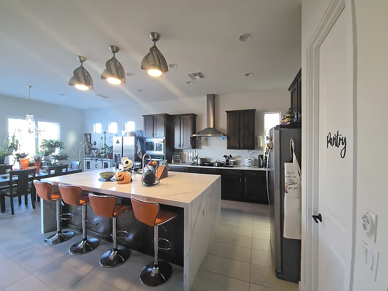 This modern kitchen features dark wood cabinets, stainless steel appliances, and a spacious white island with orange bar stools. Pendant lights, premium cookware, and playful seasonal decor create a warm, functional space perfect for cooking and entertain