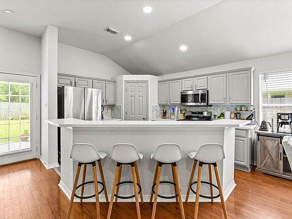 This kitchen features modern stainless steel appliances, light gray cabinetry, and a spacious island bar. It has an open layout with plenty of natural light, hardwood floors, and access to a backyard through a glass door.