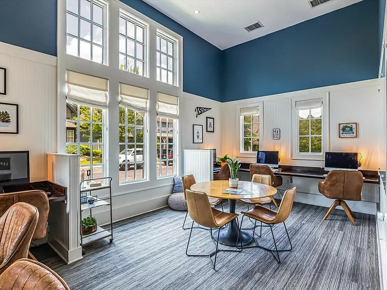 Stylish and well-lit community workspace at The Cottages of Hattiesburg featuring computer stations, a round table, and large windows with natural light, perfect for studying or remote work.