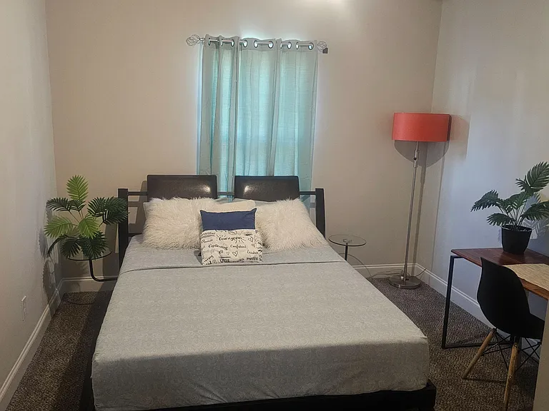 Furnished bedroom with a full bed, nightstand, and dedicated desk space, plus a window with curtains for privacy.